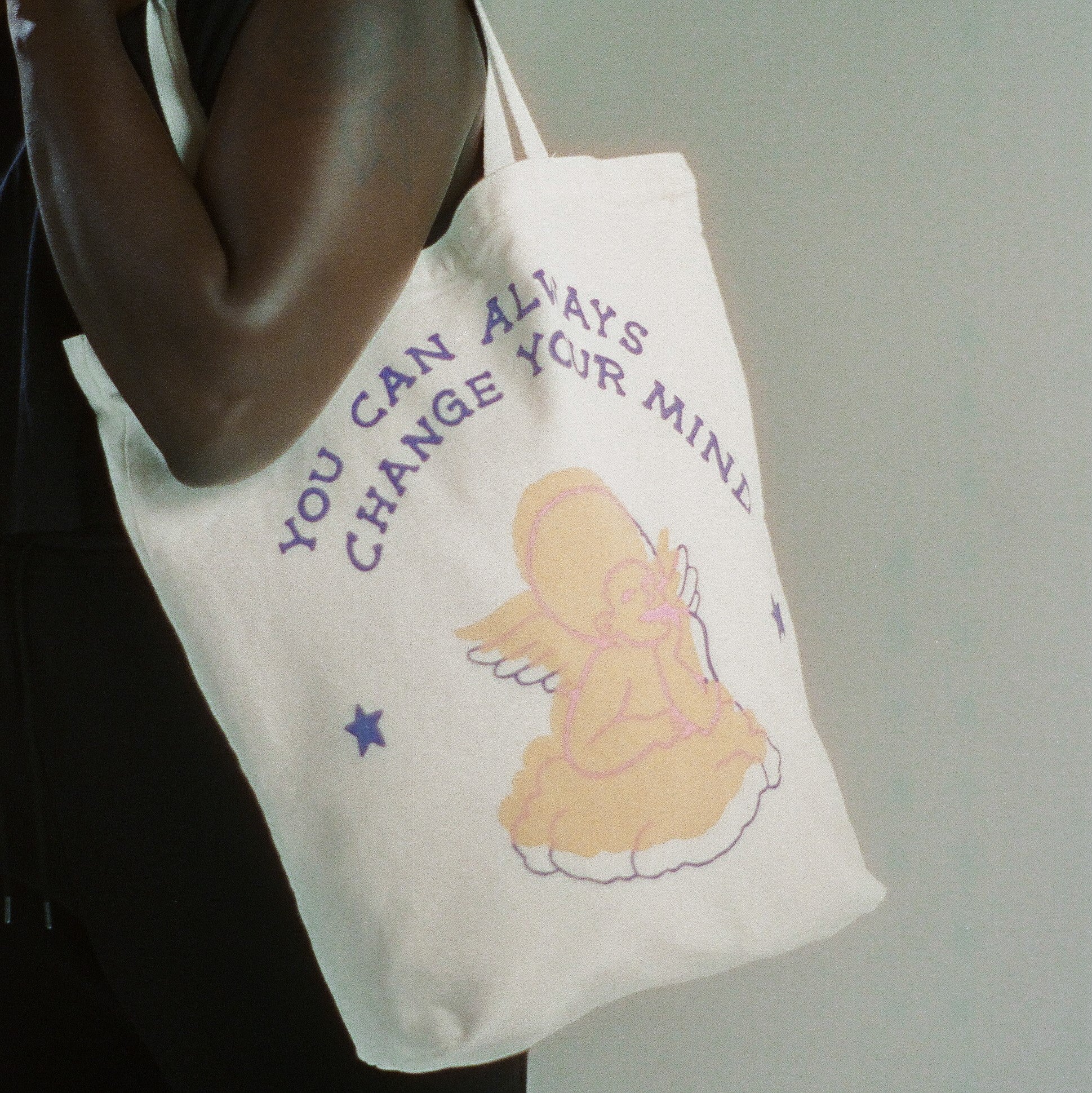 White canvas tote bag with writing that says "you can always change your mind" in dark-blue text, with a hand-drawn image of a pastel-pink angel.