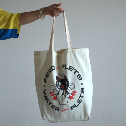 Natural canvas tote bag with text that says "feed your plets, water your plets" in black. The text surrounds a hand-drawn image of a cat in a flowerpot.