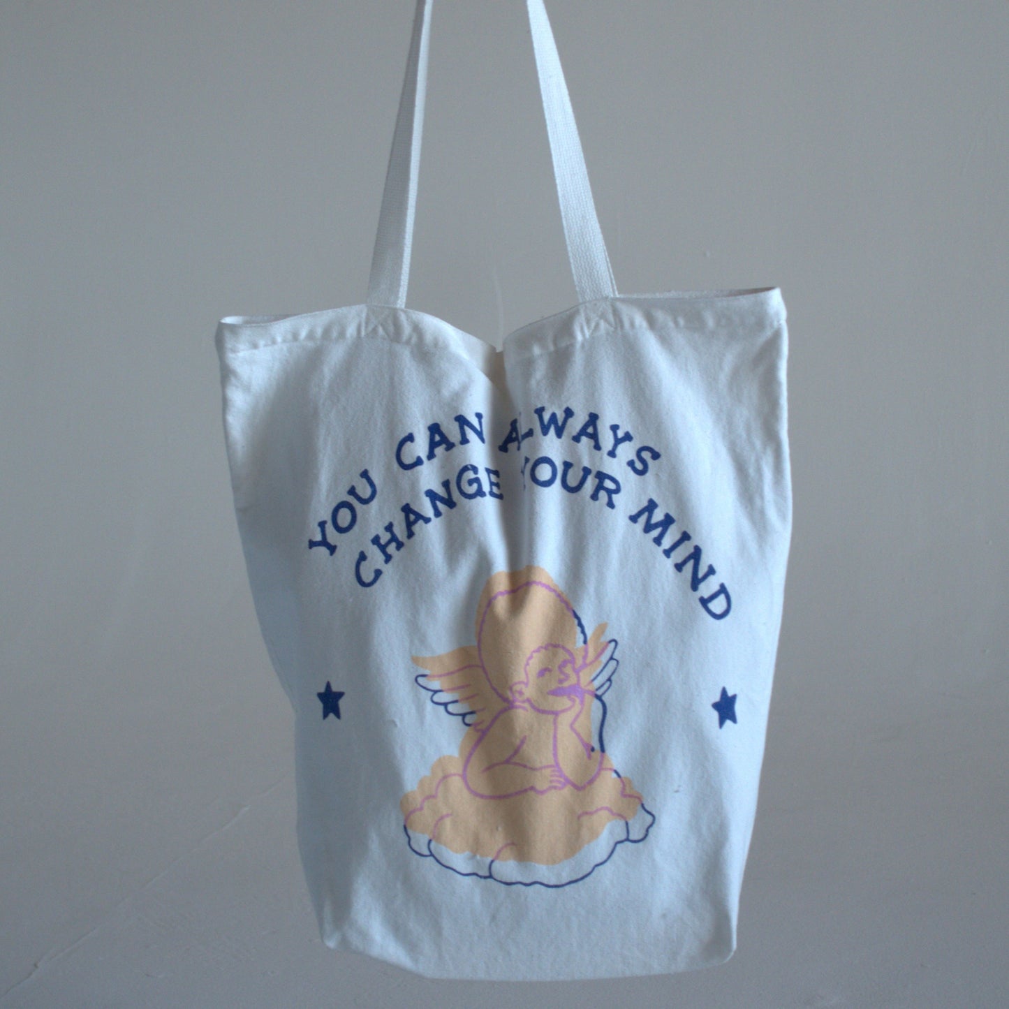 White canvas tote bag with writing that says "you can always change your mind" in dark-blue text, with a hand-drawn image of a pastel-pink angel.