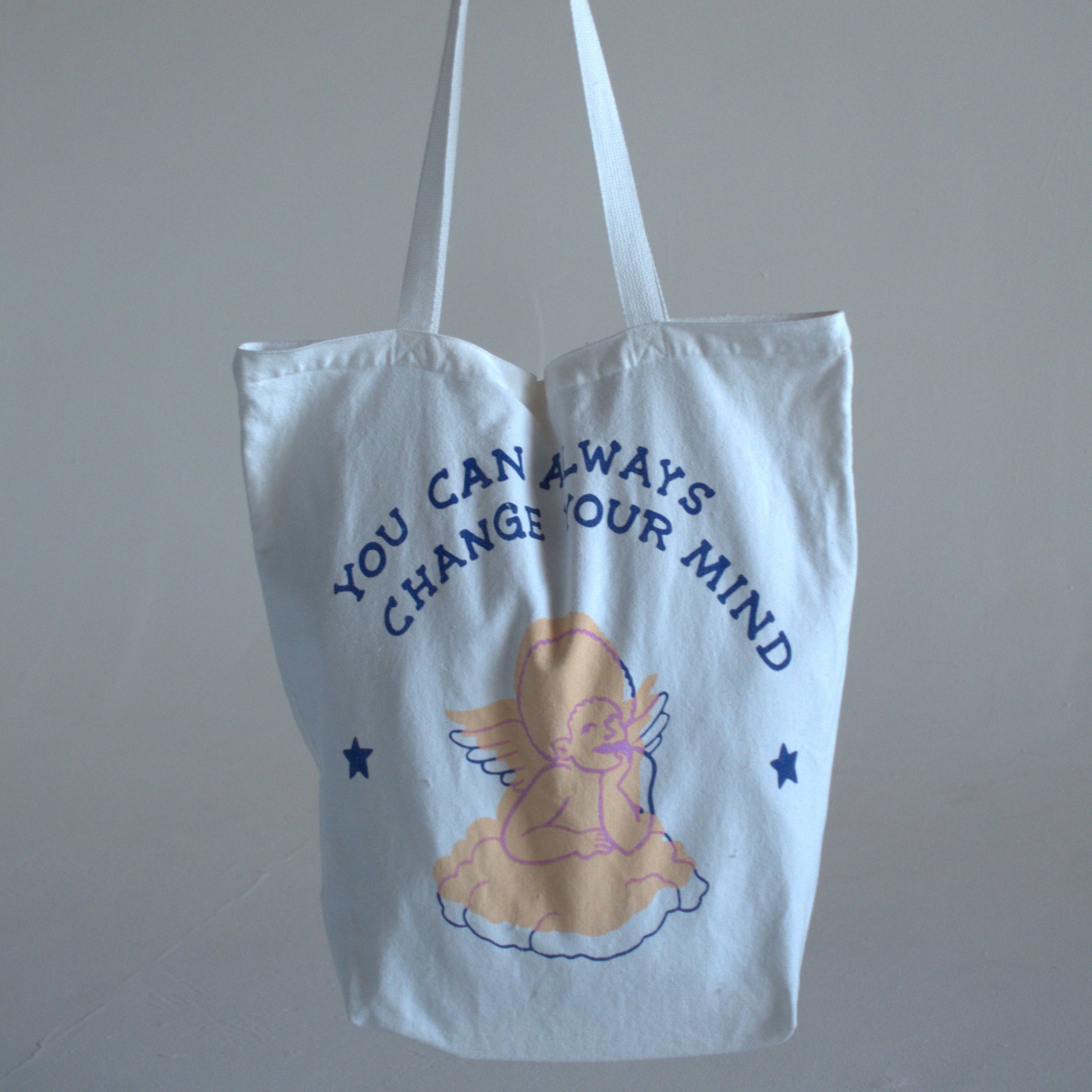 White canvas tote bag with writing that says "you can always change your mind" in dark-blue text, with a hand-drawn image of a pastel-pink angel.