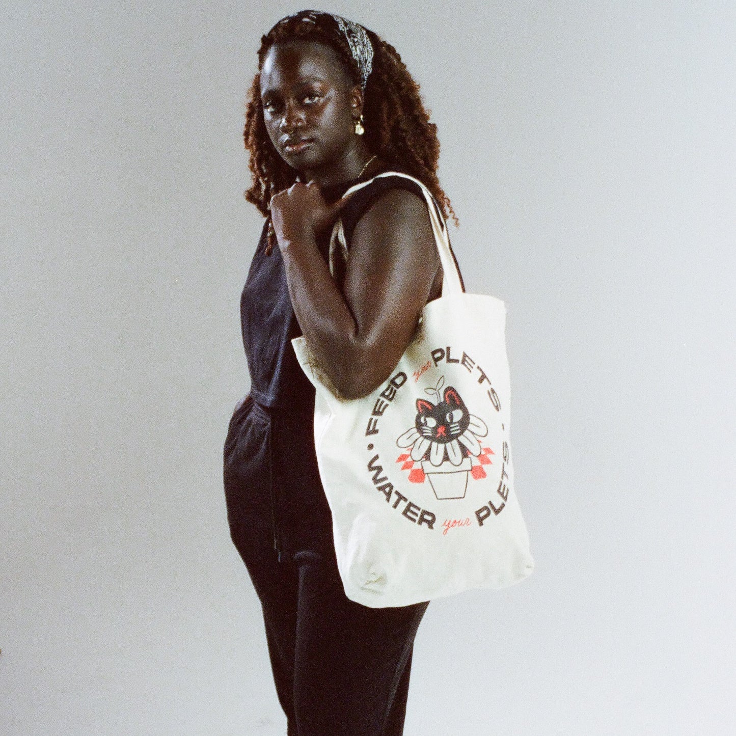 Natural canvas tote bag with text that says "feed your plets, water your plets" in black. The text surrounds a hand-drawn image of a cat in a flowerpot.